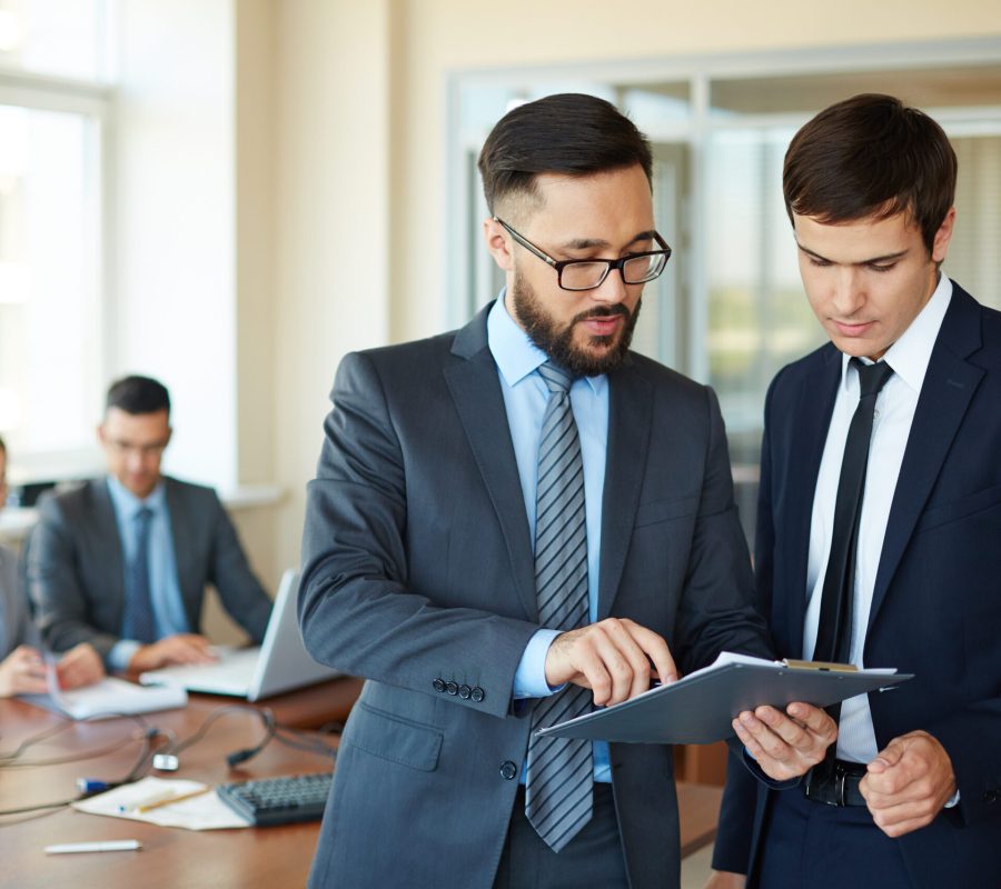 Confident businessman pointing at document while explaining his idea to his partner on background of their colleagues