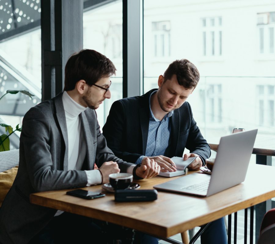 Two businessmen talking about new opportunities sitting with laptop at desk near large window, planning project, considering business offer, sharing ideas while drinking coffee together